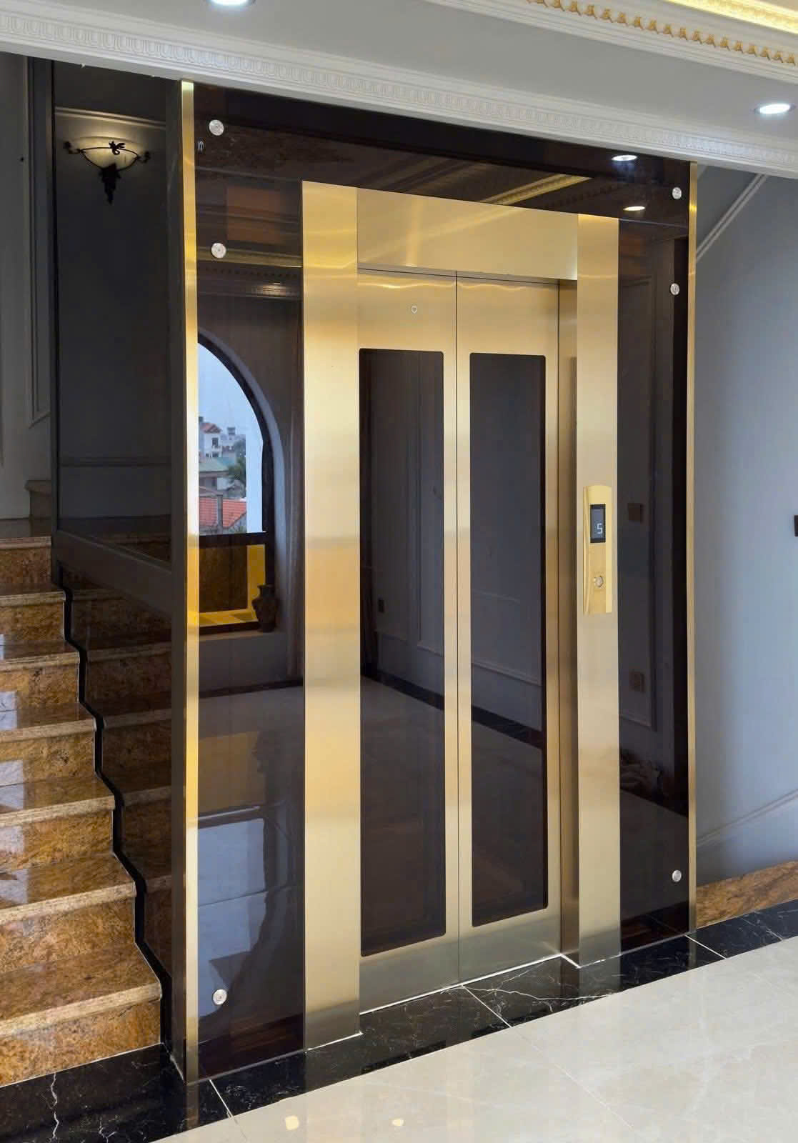 Thang máy gia đình vách kính khung inox mạ vàng sang trọng cạnh cầu thang đá marble